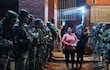 Grupo de soldados en formación, con armamento, mientras dos mujeres caminan hacia ellos, una con sudadera rosa.