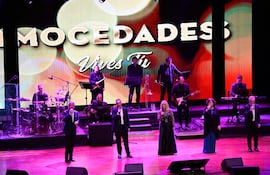 Cinco artistas, tres hombres y dos mujeres, cantan en el escenario con trajes formales y vestidos elegantes en un auditorio iluminado.