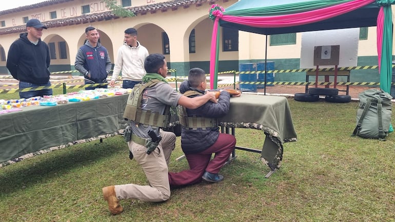 Hombre en uniforme militar ayuda a un niño en una mesa, con un ambiente de formación y un objetivo de tiro al fondo.