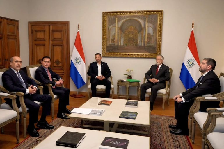 Cinco hombres en trajes formales sentados en un despacho, con banderas de Paraguay a ambos lados y documentos sobre la mesa.