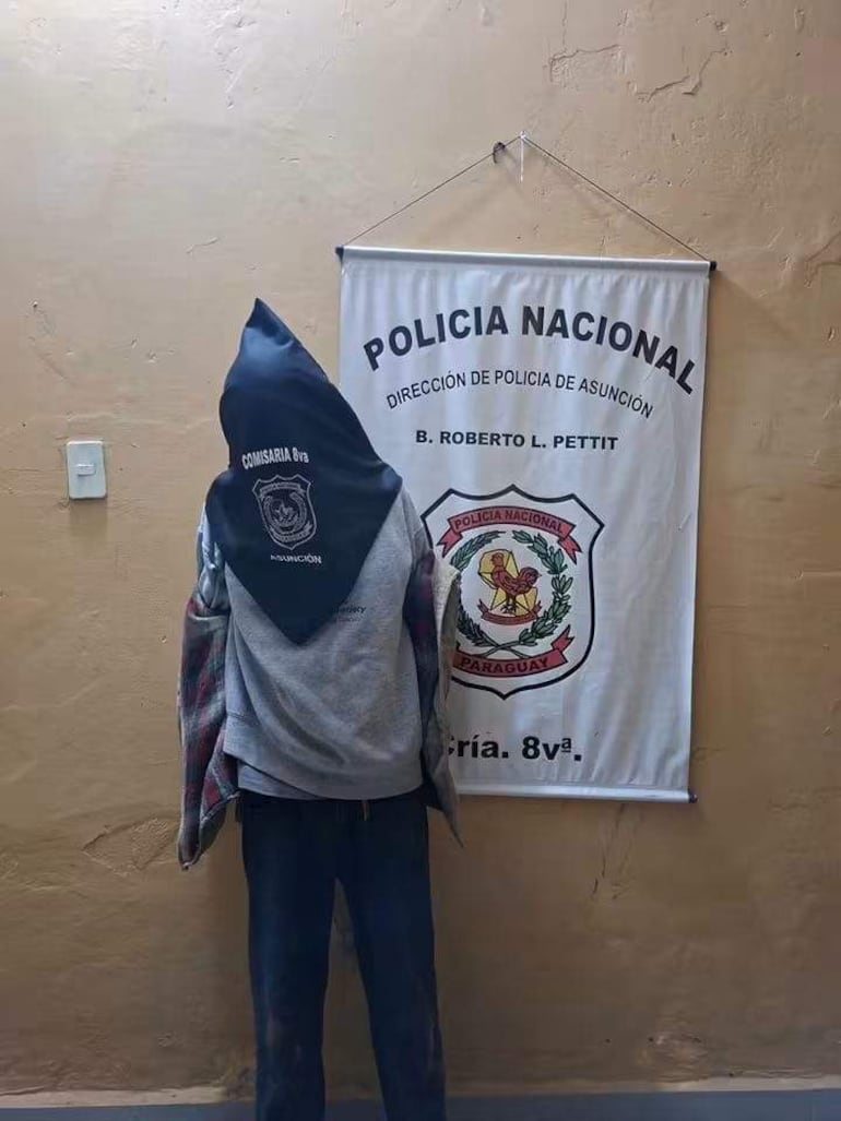 Figura humana de espaldas con pañuelo negro y abrigo de cuadros, en comisaría con banner de Policía Nacional al fondo.