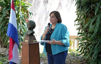 Esperanza Martínez de Portillo, con atuendo azul y blusa negra, sostiene un micrófono en un acto ceremonial, frente a un busto y una bandera de Paraguay.