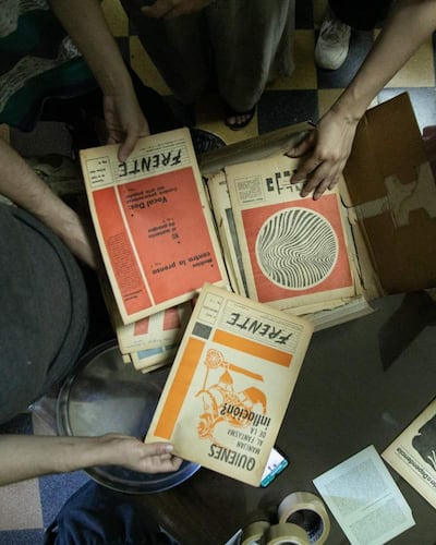 Manos revisan revistas organizadas en caja; ambiente interior con piso de baldosas de colores y objetos como cinta adhesiva y un teléfono móvil.