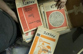 Manos revisan revistas organizadas en caja; ambiente interior con piso de baldosas de colores y objetos como cinta adhesiva y un teléfono móvil.