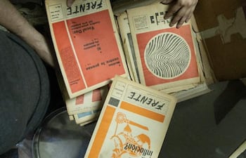 Manos revisan revistas organizadas en caja; ambiente interior con piso de baldosas de colores y objetos como cinta adhesiva y un teléfono móvil.