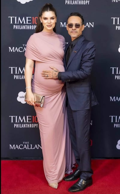 Nadia Ferreira con vestido rosa, mostrando su embarazo, y Marc Anthony en traje oscuro, ambos sonrientes en un evento elegante.
