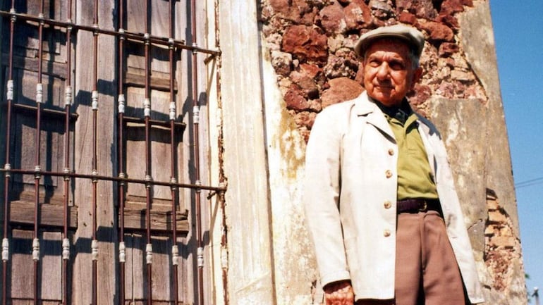 Hombre mayor con chaqueta clara y sombrero, posando frente a una pared de ladrillos expuestos.