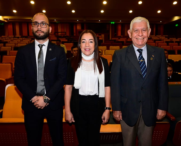 Un hombre joven con barba y gafas, una mujer con cabello lacio y chal negro, y un hombre mayor sonríen posando en un auditorio.