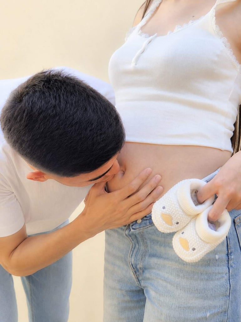 Hombre y mujer en ambiente luminoso, él se inclina hacia ella, que sostiene zapatitos de bebé con expresión significativa.