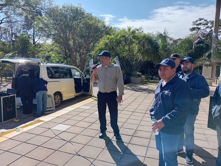 Hombre de pie con micrófono en una manifestación, dos hombres escuchando y otro preparando equipamiento en un ambiente exterior.