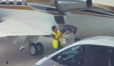 Dos hombres en el aeropuerto: uno con chaleco amarillo entrega una bolsa a otro con chaqueta clara, cerca de un avión privado.