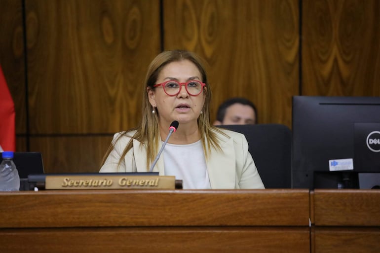 Teresa Rojas con prenda clara y gafas rojas, habla en reunión, sentada ante una mesa de deliberación con fondo de madera.