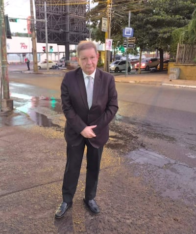 Hombre de pie en esquina con traje oscuro, camisa blanca y corbata gris, en una calle con pavimento húmedo.