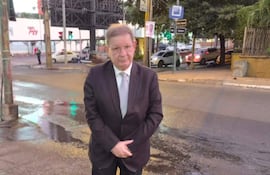 Hombre de pie en esquina con traje oscuro, camisa blanca y corbata gris, en una calle con pavimento húmedo.