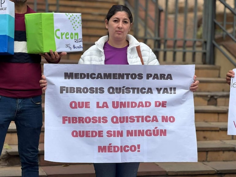 Mujer con chaqueta blanca sostiene cartel apoyando la unidad de fibrosis quística; hombre a su lado sostiene caja decorada en colores celeste y verde.