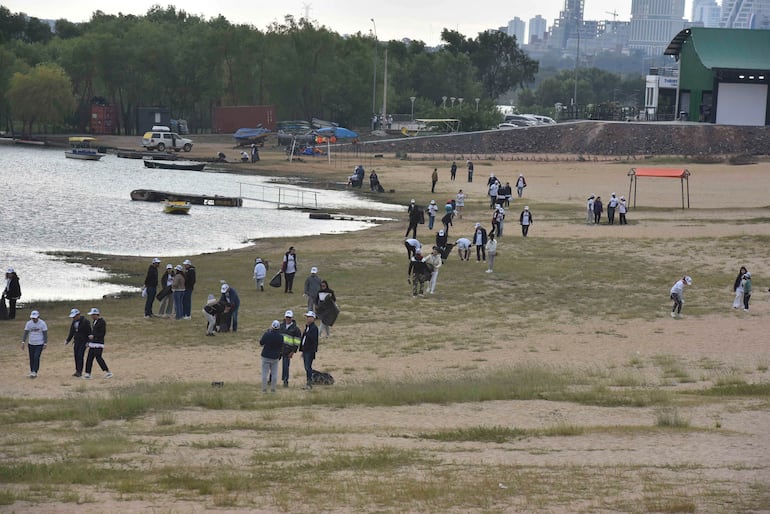 Grupo de personas con camisetas blancas y gorras limpiando la Costanera, cerca de botes y un entorno urbano.