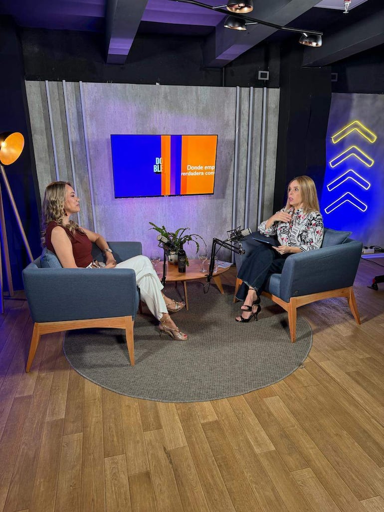 Laura Torres con blusa blanca y Belén Bogado con vestido marrón conversan en sillones azules, rodeadas de decoraciones en un estudio de grabación.