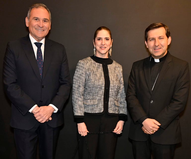 Hombre sonriente en traje oscuro, mujer en chaqueta de diseño en tonos gris y negro, y hombre en sotana negra en evento elegante.