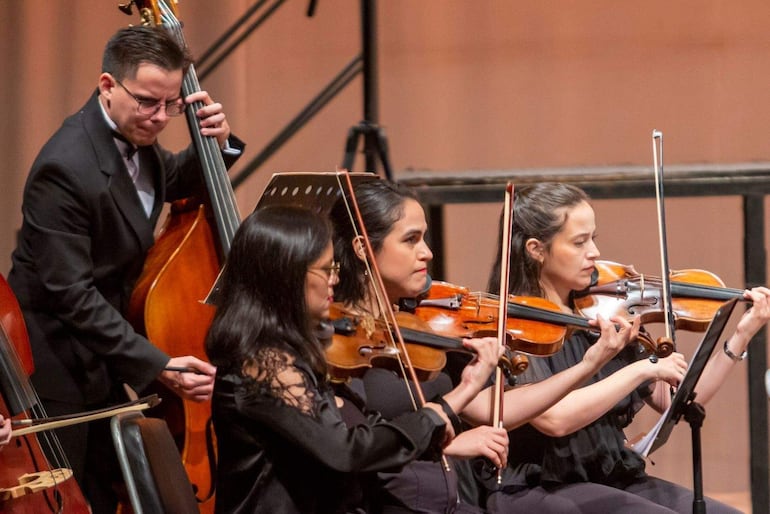 Músico con contrabajo en traje negro, flanqueado por tres mujeres violinistas vestidas de negro en un escenario formal.