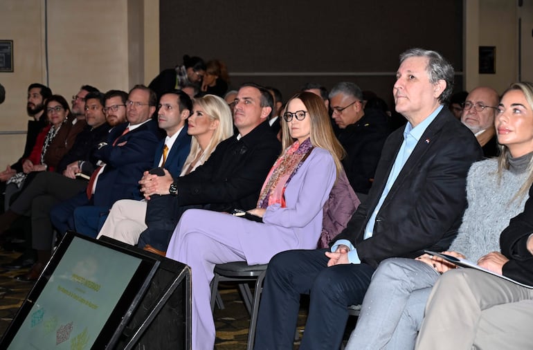 Soledad Núñez con chaqueta roja y Rafael Filizzola en auditorio, ambos atentos durante la presentación en evento formal interno.