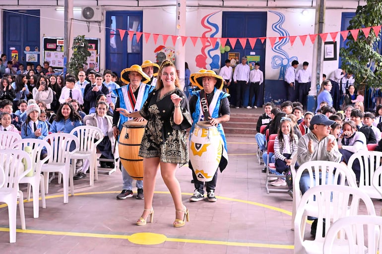 Mujer baila con vestido negro y detalles dorados, rodeada de músicos con instrumentos de percusión y estudiantes animados al fondo.