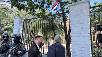 Dos hombres conversan, uno con gafas y traje negro, otro con saco azul. Agentes de seguridad armados los custodian en un entorno al aire libre.