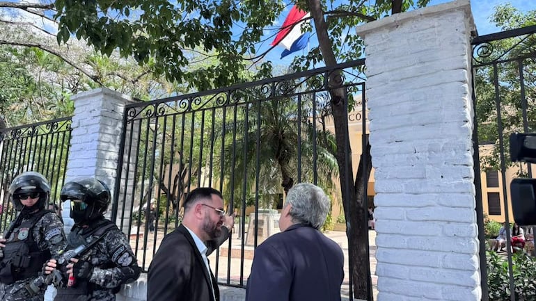 Dos hombres conversan, uno con gafas y traje negro, otro con saco azul. Agentes de seguridad armados los custodian en un entorno al aire libre.