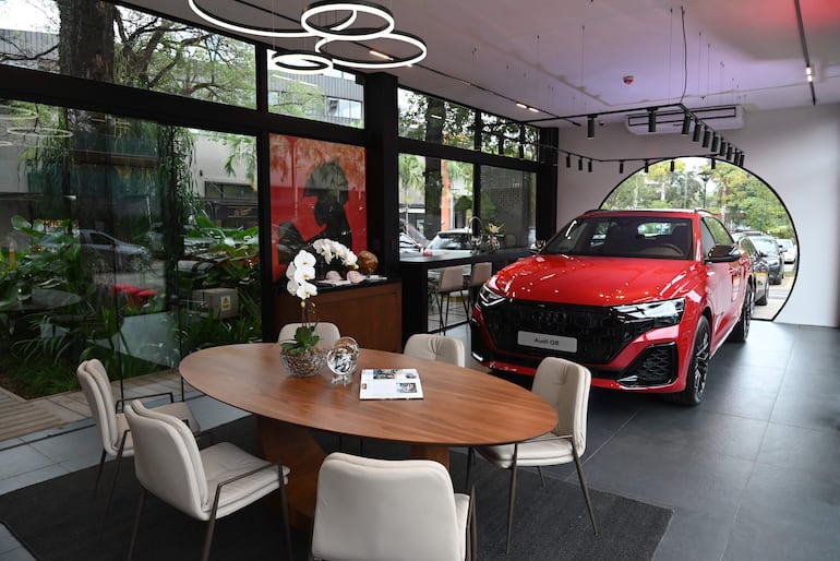 Audi Q8 rojo brillante estacionado en sala moderna con mesas y sillas blancas, decoraciones elegantes y vegetación al exterior.