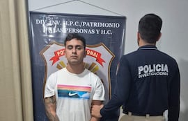 Hombre con camiseta blanca y jeans esposado, custodiado por un agente con chaqueta de Policía Investigaciones en estación policial.