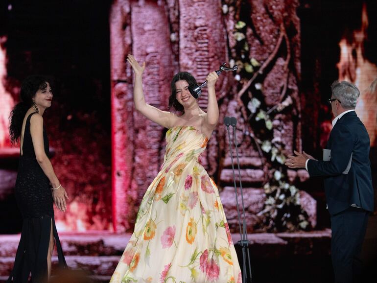 Mujer con vestido largo de flores sostiene un trofeo, rodeada de una mujer sonriente y un hombre aplaudiendo.