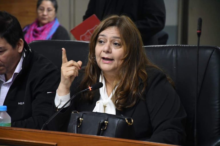 Rosanna Rolón en blazer negro y blusa blanca, señalamientos enfáticos en una reunión formal, con hombre y mujer escuchando atentamente.