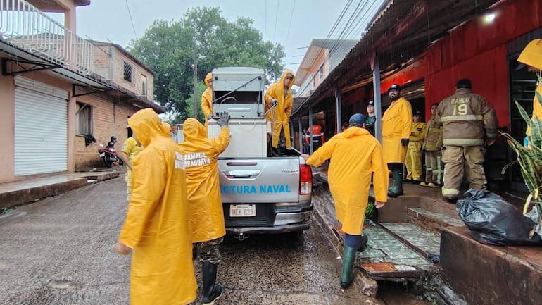 Seis trabajadores con impermeables amarillos colaboran en un entorno urbano lluvioso, cerca de un vehículo de la Prefectura Naval.