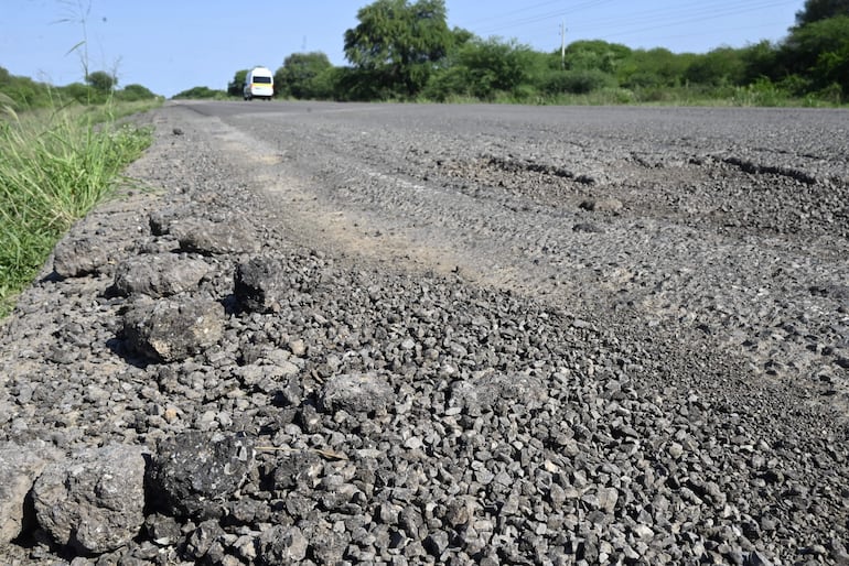 Carretera deteriorada con piedras y grava, vehículo blanco en movimiento y vegetación densa a los lados.
