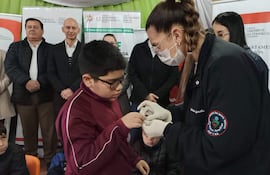 Niño en camiseta borgoña recibe tratamiento de salud por profesional con guantes, observados por adultos en un centro de salud.