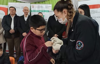 Niño en camiseta borgoña recibe tratamiento de salud por profesional con guantes, observados por adultos en un centro de salud.