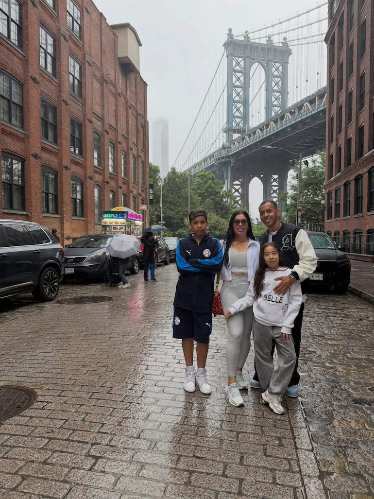 Alejandro Romero sonríe junto a dos niños vestidos de un equipo de fútbol y una mujer en camiseta clara, bajo una lluvia ligera.