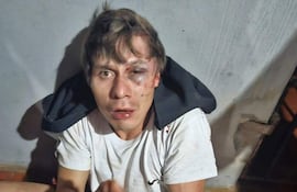 Hombre sentado en el suelo, con hematoma en el ojo y sangre en el labio, esposado y con camiseta blanca.