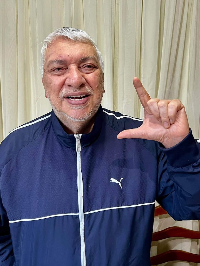 Fernando Lugo, canoso y sonriente, viste chaqueta deportiva azul mientras hace el gesto de la 'L' con su mano.