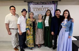 Siete adultos sonrientes en un evento, variando en vestimenta: hombre con camisa de cuadros, mujeres con abrigo colorido y chaqueta de mezclilla.