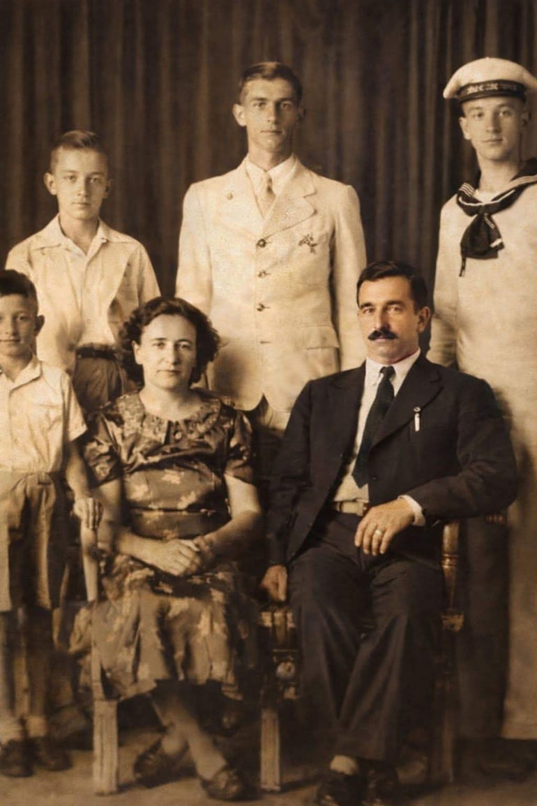 Mujer con vestido floral sentada junto a hombre con bigote, rodeados de cuatro hombres en trajes formales, fondo oscuro.