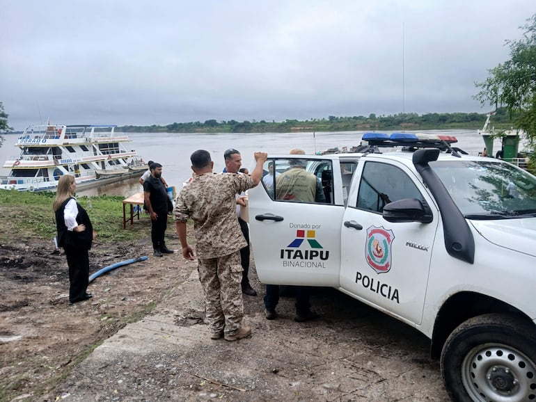 Un hombre en uniforme camuflado discute con otros, mientras un barco de pasajeros y un vehículo policial están presentes en un día nublado.