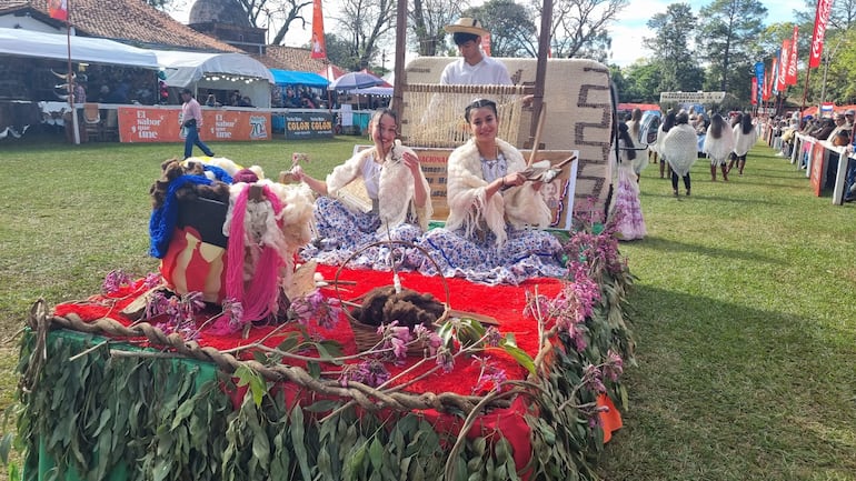 Dos mujeres en vestimenta tradicional con faldas largas y chaquetas de lana. Un hombre de pie realiza labores de textil en un ambiente festivo.