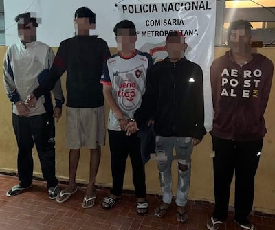 Cinco hombres esposados, algunos con chaquetas y pantalones oscuros, en comisaría con cartel de 'Policía Nacional' de fondo.