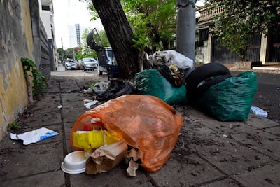 Acera cubierta de sacos de basura negra y verde, con un árbol al fondo y vehículos estacionados en el camino.