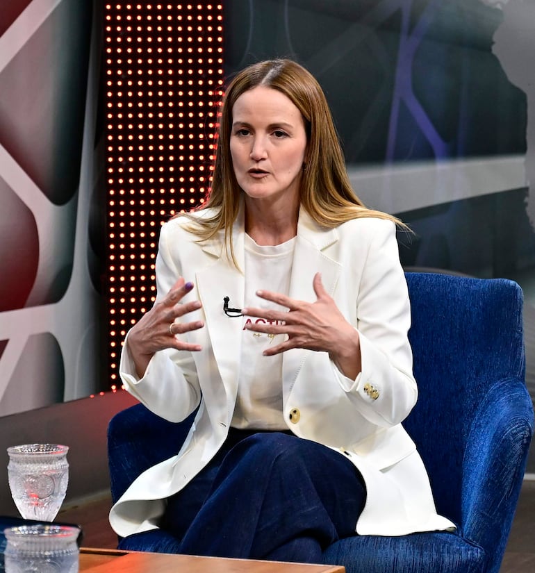 Soledad Núñez sentada en sillón azul, con chaqueta blanca y pantalones oscuros, gesticulando mientras habla en un set televisivo.