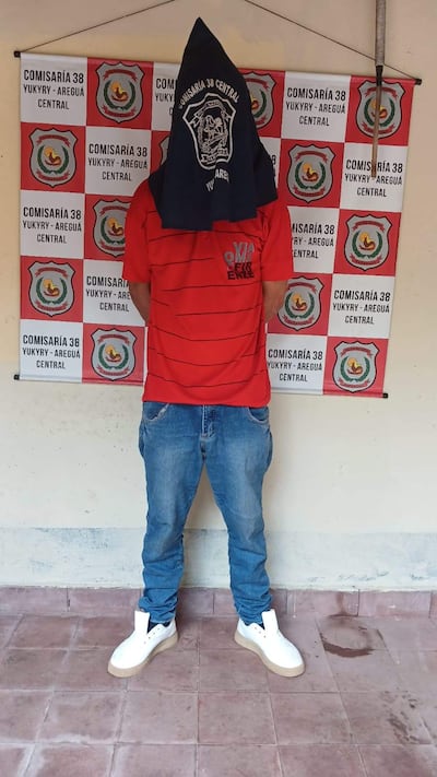 Persona de espaldas con camiseta roja y pantalones de mezclilla, cubriendo su cabeza con una toalla, frente a la Comisaría 38 de Yukyry.