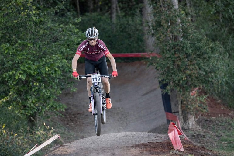 Ciclista en camiseta burdeos y pantalones cortos negros realizando un salto sobre una rampa de tierra en un bosque.
