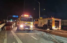 Camiones de bomberos amarillos y claros iluminan la escena nocturna en Pedrozo, mientras sombras se asoman al fondo.