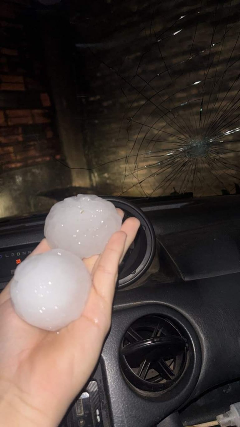 Mano sostiene grandes bolas de granizo en un vehículo, con ventana agrietada y tablero visible.