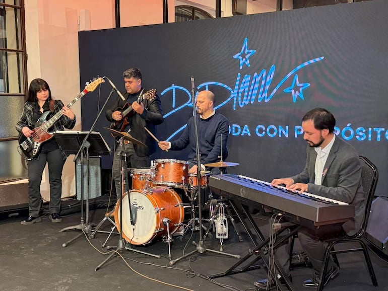 Músicos en escenario: mujer con bajo, hombre con guitarra, batería y teclado, iluminados por luces suaves en evento musical.